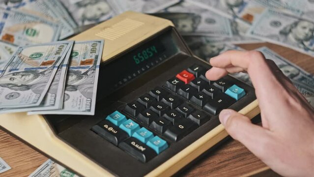 Counting on an old vintage calculator surrounded by dollar bills, close-up. Male hand press keyboard buttons on an old retro calculating machine. Counts finances, currency, savings, financial bills 4K