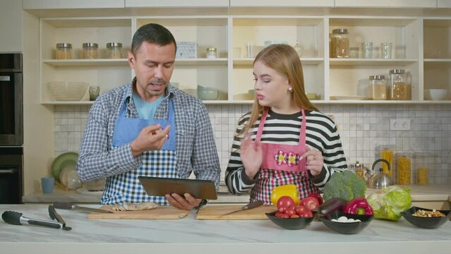 Attractive hearing impaired family in aprons preparing for cooking tasty dinner, checking necessary food ingredients on digital tablet and talking with sign language in domestic kitchen.