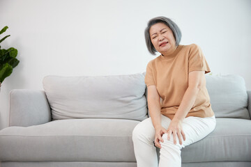 senior woman suffering from knee ache on sofa - obrazy, fototapety, plakaty
