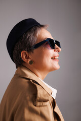 Smiling and stylish mature woman in sunglasses, beret and classic trench coat isolated on grey