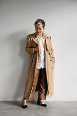 Stylish and elegant gray haired woman in trench coat, eyeglasses and heels standing near wall 