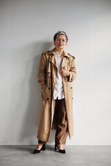 Confident and elegant mature woman in trench coat and eyeglasses standing near grey wall 