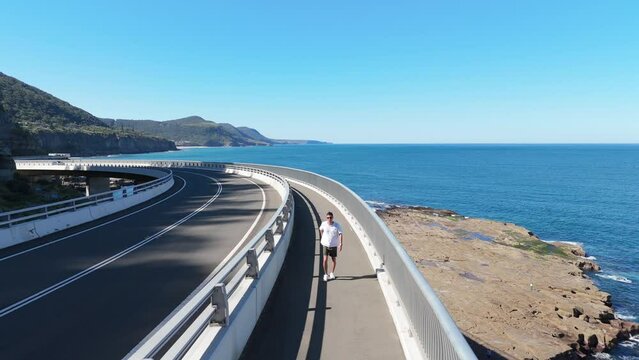 young man walking, jogging and running along the sea cliff bridge as vehicles pass by him. Below you can see the sea and the top of the bridge avoiding the cliff between the rocks and the sea