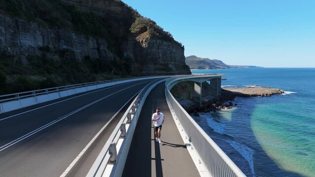 young man walking, jogging and running along the sea cliff bridge as vehicles pass by him. Below you can see the sea and the top of the bridge avoiding the cliff between the rocks and the sea