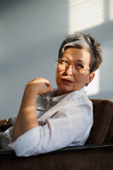 Portrait of elegant mature woman in eyeglasses and white shirt sitting on armchair on grey with...