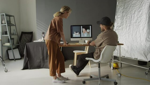 Full shot of young Caucasian female photographer walking up to desk of male colleague, looking together at photos of commercial product samples on computer screen, and discussing best angle