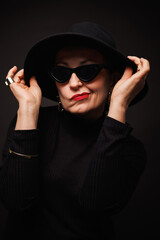 Portrait of stylish mature woman with red lips posing in sunglasses and hat isolated on black 