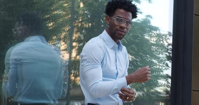 Young handsome African American man in blue shirt, stylish glasses and earrings stands near building in city, straightens his clothes and looks at camera, exuding confidence and charm.