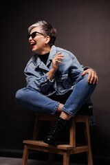 Cheerful and trendy mature woman in sunglasses and denim jacket posing on black background