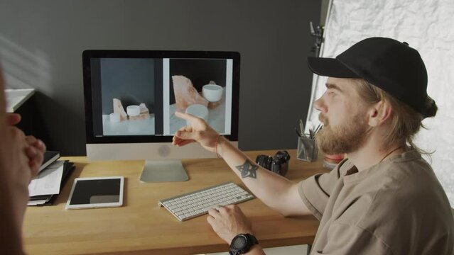 Medium close-up shot of Caucasian male graphic designer sitting at desk in front of computer, talking to unrecognizable female colleague, demonstrating best ways to shoot product samples on monitor