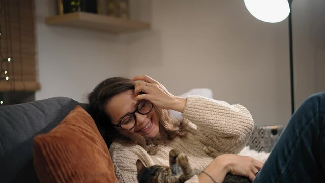 A young Caucasian woman playing with a cute grey cat sitting on the couch at home on a winter evening