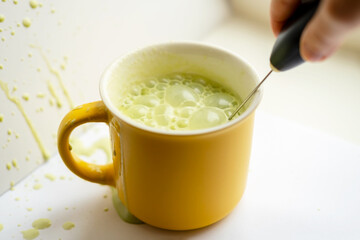 Woman hand is brewing and mixing matcha green latte tea in yellow mug. Isolated on white. Beating with a whisk