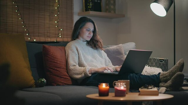 A young Caucasian woman working on a laptop while sitting on the sofa on a Christmas winter evening