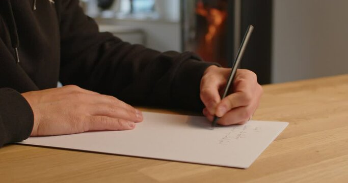 Caucasian man writing with left hand. Medium close up shot of writing with a pencil on a piece of paper. Cozy fireplace with fire in the background.