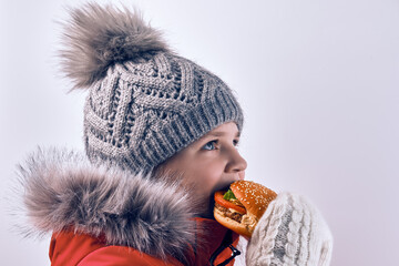 A 10-11 year old girl eats a hamburger in winter clothes, against a light background in the studio.
