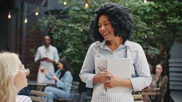 View of pretty waitress taking order of pretty female client on pastry shop. Multi-ethnic women communicating with each other. Beautiful female worker recommending to choose tasty food.