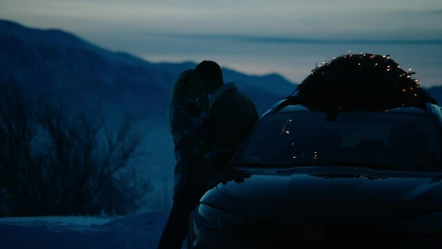 Silhouette of a couple kissing near the car with lit Christmas tree tied to the roof, winter mountains in the background