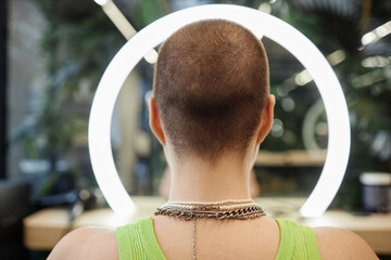 Back view of unrecognizable young woman with buzzcut looking at ring light in beauty salon taking...