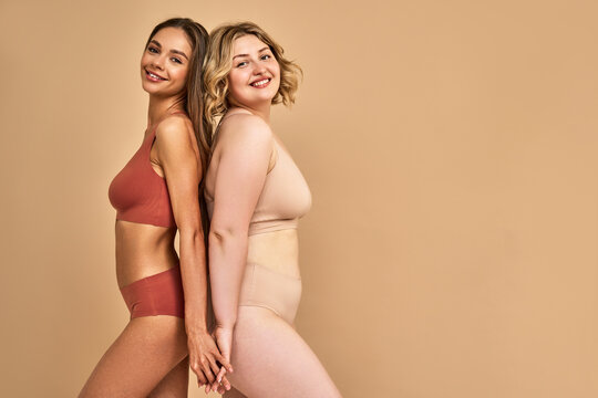 Different body types. Young skinny lady and plus size woman standing back to back and holding hands over beige background. Two females posing in studio in red and beige sports underwear. Copy space.