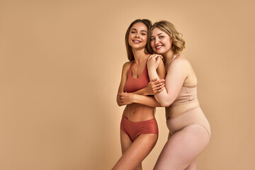 Female beauty. Positive ladies with blond and dark hair posing in gentle hugs over beige studio background. Two ladies with different skin color and body size expressing support and acceptance.