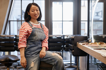 Portrait of smiling young Asian woman as hairstylist looking at camera posing at beauty salon workplace and wearing jeans overalls, copy space