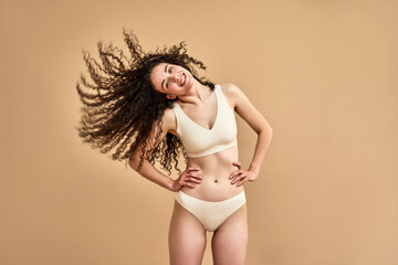 Lady's allure. Positive caucasian woman playing with her long wavy locks while standing in studio and smiling at camera. Attractive young female with slim body shape wearing comfy skin color lingerie.
