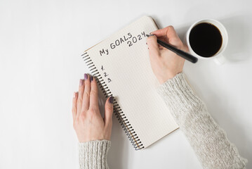 Female hands writing My Goals 2024 in a notebook. Mug of coffee on the table, top view