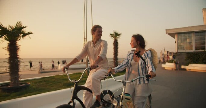 Front view of a guy and a girl riding bicycles and looking at each other on the beach on which palm trees grow near the sea in summer at dawn. Bike ride on the beach in the morning
