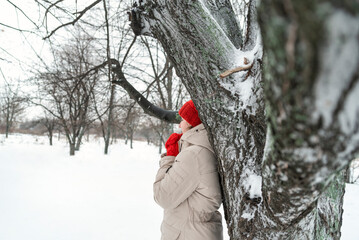 Girl stands leaning her back against tree in snowy forest. Woman enjoys beauty of winter landscape. Trees covered in snow