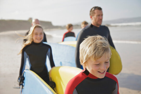 Boy and girl carrying surfboards on a beach followed by people
