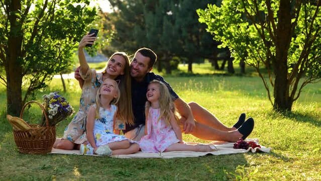 Happy family having picnic sitting on blanket in park on sunny day and taking selfie photos on smartphone. Young cheerful parents taking pictures with kids on mobile phone outdoor. Real time.