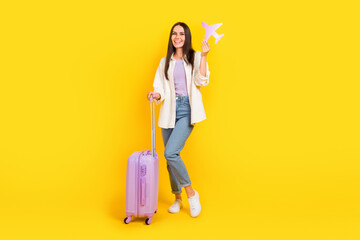 Full length photo of good mood woman dressed white jacket hold luggagge paper plane go on vacation isolated on yellow color background