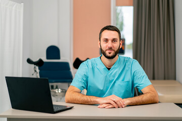 Portrait of a serious male doctor in uniform sitting at his desk with a laptop in a medical waiting room and looking at camera
