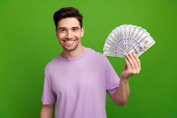 Photo of successful man with brunet hair dressed purple t-shirt presenting bunch of dollars in arm isolated on green color background