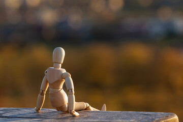 A wooden mannequin sits on a bench and looks at an autumn blurred landscape. The concept of loneliness,