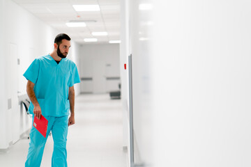 A tall male doctor with a beard walks along the hospital corridor with folder of documents and...