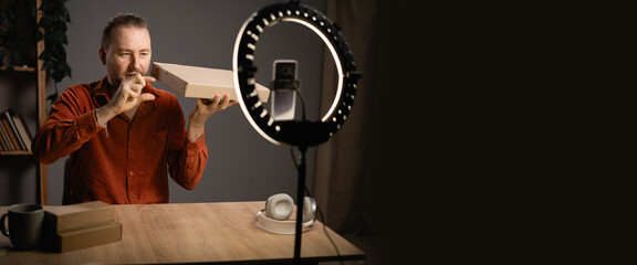 Millennial man recording unboxing video sitting at home in front of smartphone camera and ring lamp. Blogging and review product concept