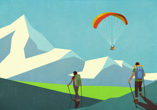 Couple hiking below paraglider in sunny mountains
