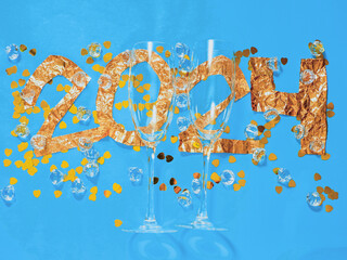2024 hand made sign made from golden color foil, two tall elegant champagne glasses and heart shaped confetti on blue background. New Year celebrations concept.