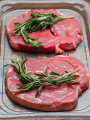 Juicy uncooked lamb leg steak with green fresh rosemary on plastic tray on wooden table. Uncooked high quality meat product with rich taste and vivid color.