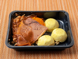 Classic beef dinner meal with slice of meat, mashed potatoes, gravy and turnip in dark plastic container on bamboo placemat background. Microwaveable food for take away or business lunch.