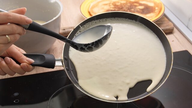 Pouring Batter onto Pan: A person's hand holds a ladle, guiding a smooth flow of batter onto a hot pan. Adjacent to this, a cooked crepe rests on a wooden board. Making pancake. Cooking food. 