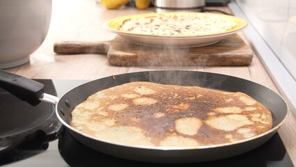 Cooking a pancake in a frying pan with steam. Next to the stove, there's a freshly cooked pancake on a wooden board. The setting appears to be a modern kitchen, ready for preparing food.
