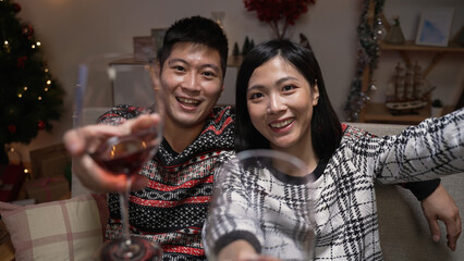 mobile cam view of a smiling Japanese asian couple drinking a Christmas toast to their friends during an online virtual chat in the living room at home. - obrazy, fototapety, plakaty