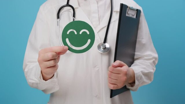 Close up of young doctor woman in white uniform and stethoscope holding green happy smiley, isolated on blue background, satisfaction survey, child wellness, world mental health day, Compliment Day
