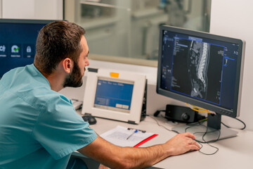 Concentrated male physician sits at a computer monitor in a room for describing MRI images and monitoring patient's condition - obrazy, fototapety, plakaty
