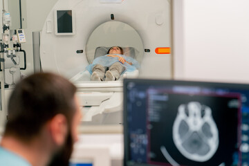 A radiologist sits at a table behind a computer monitor and examines a magnetic resonance imaging image during an examination of patient - obrazy, fototapety, plakaty