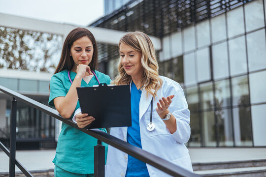 Two mid adult female healthcare professionals walk and talk together. Female Medical Staff In White Coats And Scrubs Use Clipboard In Informal Meeting In Hospital