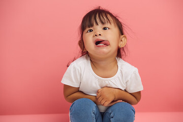 Funny little child girl capriciously shows his tongue, sits on the floor on a pink isolated background, close up portrait.