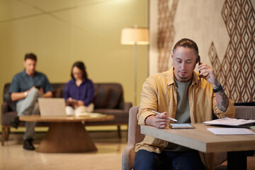 Portrait of startupper talking on phone with cofounder when checking report on tablet computer